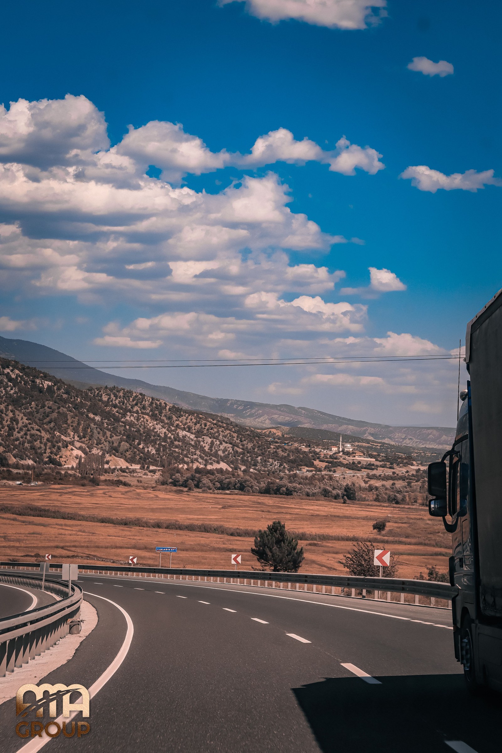 Camion pe o autostradă care traversează un peisaj montan arid, sub cer albastru